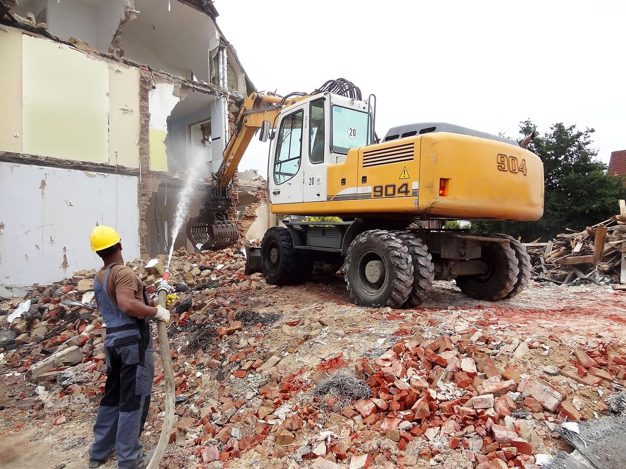 Excavator tearing down a building while a worker wets the rubble